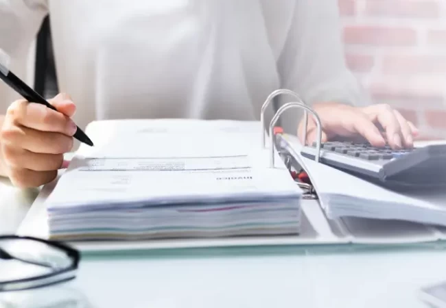 A person working at a desk with documents, using a calculator and holding a pen.