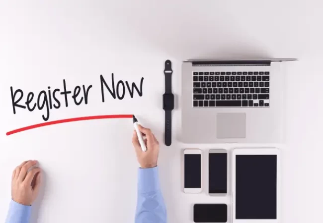 A person's hands are seen at a desk, holding a marker and writing "register now" above a neatly arranged set of devices that includes a laptop, a tablet, a smartphone, and a smart