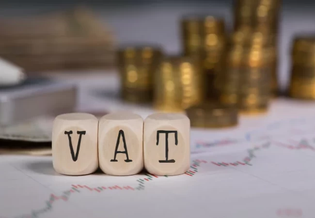 Three wooden cubes with letters spelling "vat" in front of a blurred background with stacks of coins and financial documents