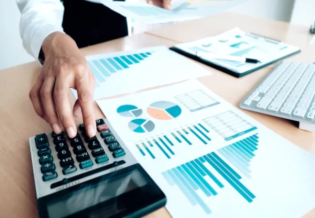 A person is using a calculator next to several financial charts and graphs on a desk with a computer keyboard in the background