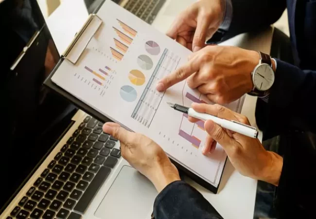 Two individuals in a business setting are reviewing and discussing a chart and graph on a document, with a laptop and additional papers adjacent to them