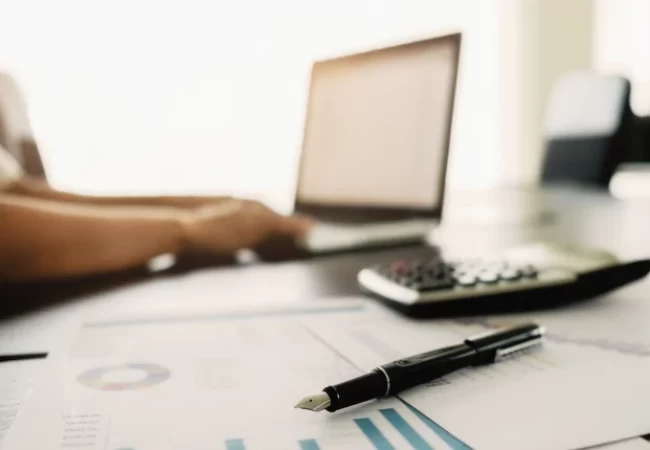 Person at a workstation with a laptop, calculator, and documents including graphs, indicating financial or business analysis